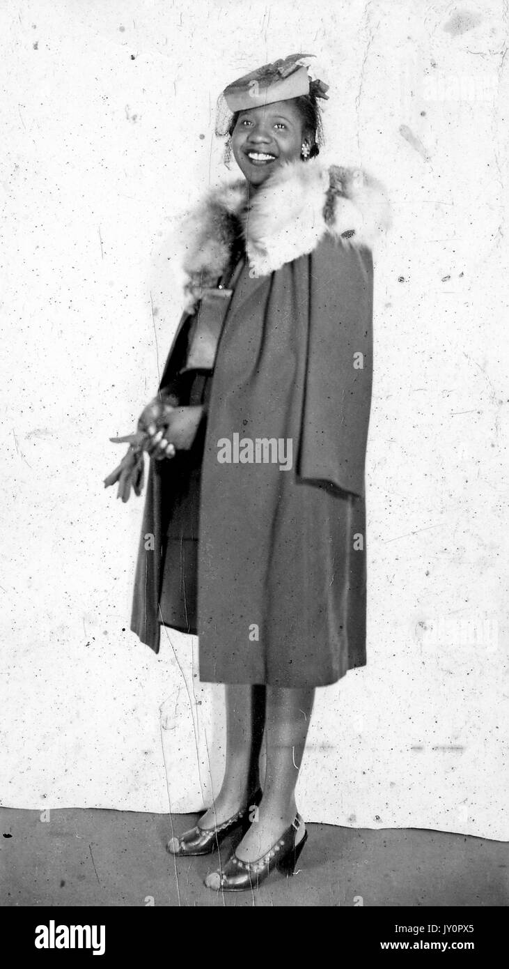 Portrait debout, jeune femme afro-américaine, portant une paire de gants sombres, portant un pardessus sombre avec col en fourrure, bijoux et chapeau, expression souriante, 1920. Banque D'Images