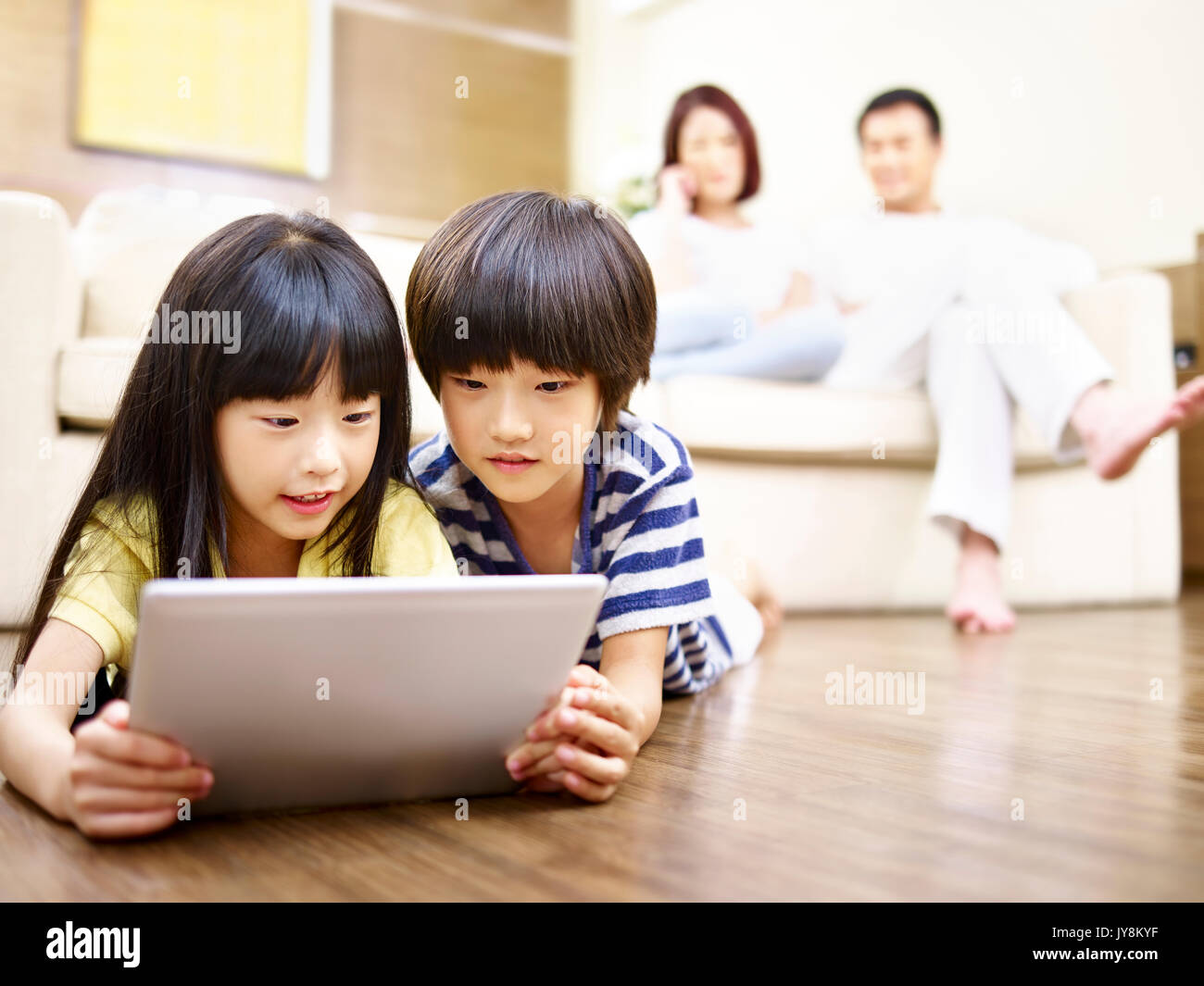 Deux enfants asiatiques lying on floor playing video game using digital tablet while parents regardant en arrière-plan. Banque D'Images