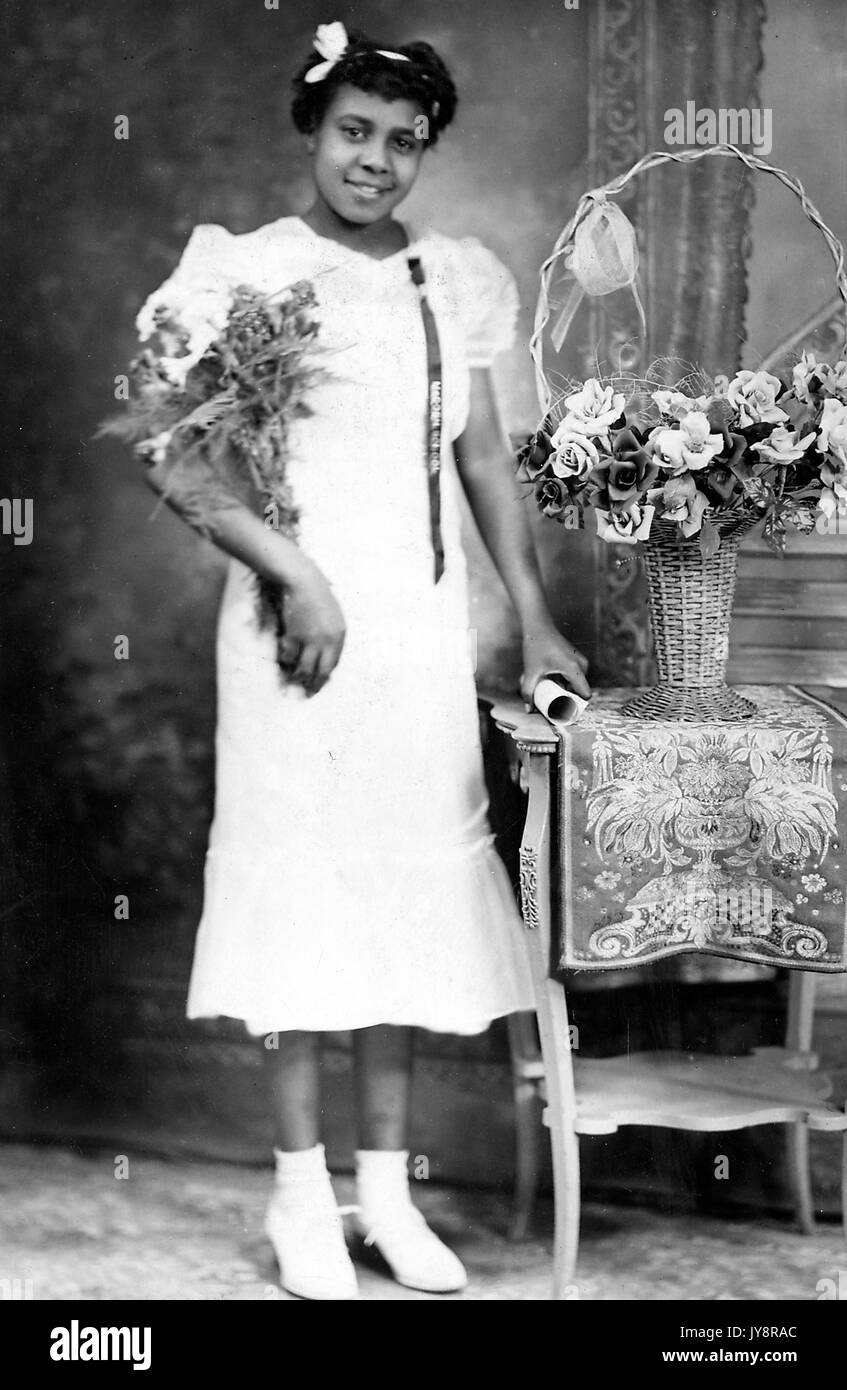 Portrait debout de jeune femme afro-américaine souriante, portant une robe légère et des chaussures avec une fleur dans ses cheveux, portant des fleurs, sa main gauche, qui détient un diplôme, repose sur une table avec une nappe et un panier de fleurs, 1915. Banque D'Images