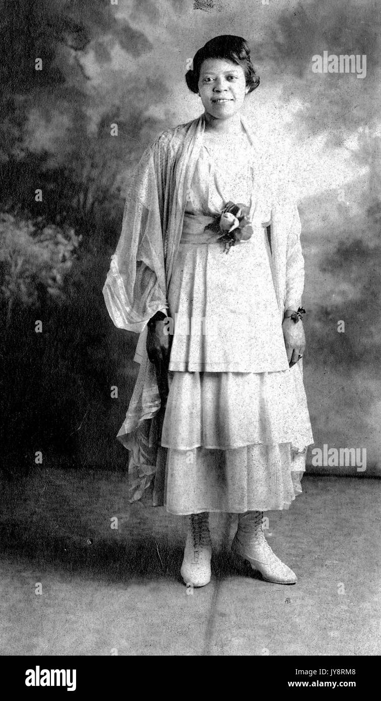 Permanent pleine longueur portrait of smiling young african american woman in front of painted background, vêtu d'une robe légère et chaussent des bottes, 1915. Banque D'Images