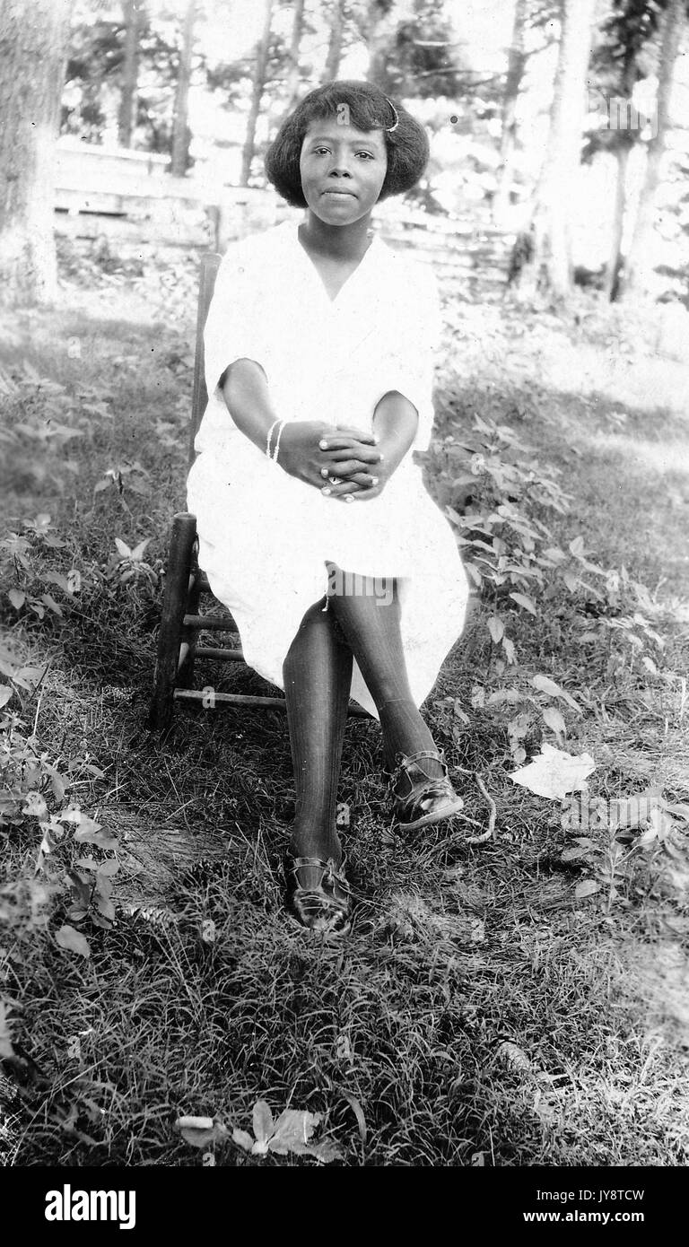 Young african american woman qui pose pour un portrait dans un cadre boisé, compensation assis jambes croisées sur une chaise et vêtue d'une robe blanche, avec une expression du visage, 1944. Banque D'Images