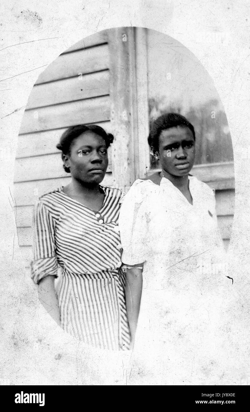 Trois quarts oval portrait de deux jeunes femmes afro-américaines, près d'une maison, l'autre sur la gauche dans une robe rayée, l'autre dans une robe blanche, à la fois avec les expressions du visage tendus, 1920. Banque D'Images