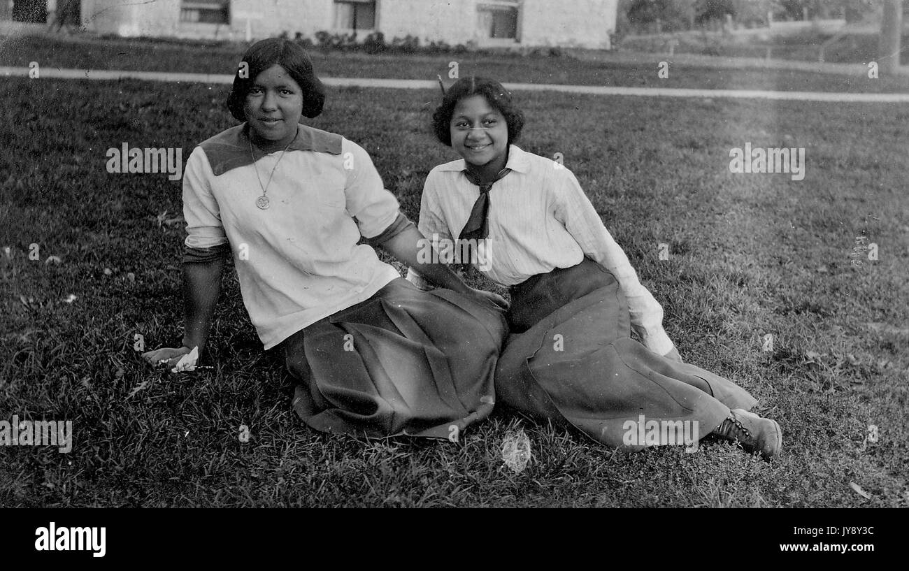 Portrait en pied assis, deux femmes afro-américaines, âgées de 8 à 18 ans, une à droite plus jeune qu'une à gauche, assis à l'extérieur dans l'herbe, portant de longues jupes avec des hauts à manches longues, expressions souriantes, 1915. Banque D'Images