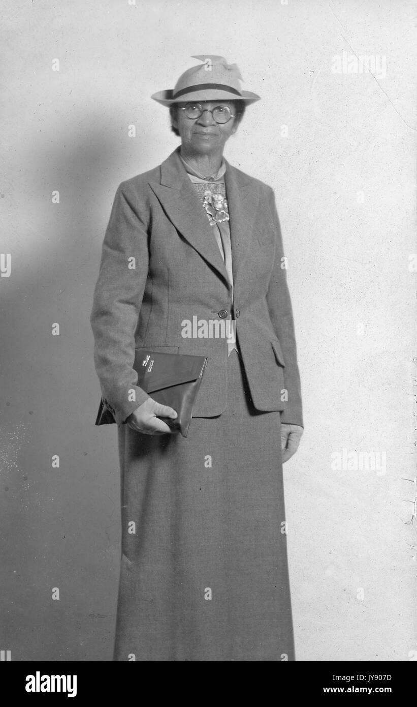 Portrait debout de trois quarts de longueur de femme afro-américaine âgée, portant une jupe sombre, un chemisier, une veste, des gants, des lunettes et un chapeau, pochette de transport, expression neutre, 1925. Banque D'Images