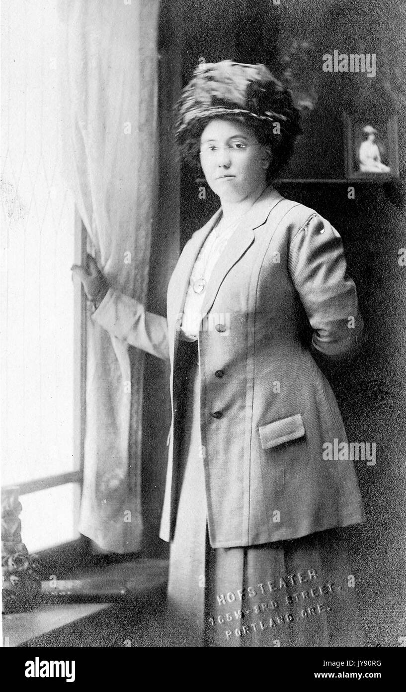 Portrait d'une femme afro-américaine debout à côté d'une fenêtre, portant un chapeau, des lunettes et une veste et une jupe en deux pièces, avec une photo encadrée d'un portrait d'une femme derrière elle, Hofsteater, 165 W - 3rd Street, Portland, Oregon, 1915. Banque D'Images