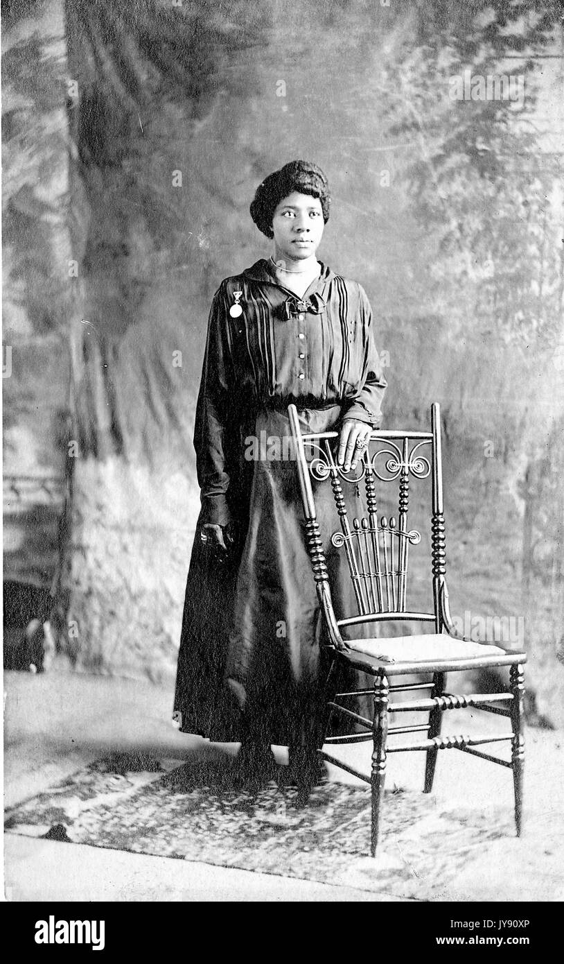 African-American Woman, portrait complet du corps, debout derrière une chaise, la main posée sur une chaise, vide d'expression, 1915. Banque D'Images