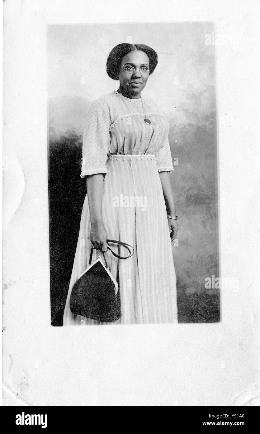 Portrait debout de trois quarts de longueur de femme afro-américaine mature, expression neutre, portant un sac à main, portant un collier, un bracelet et une robe à motifs de couleur claire, 1915. Banque D'Images