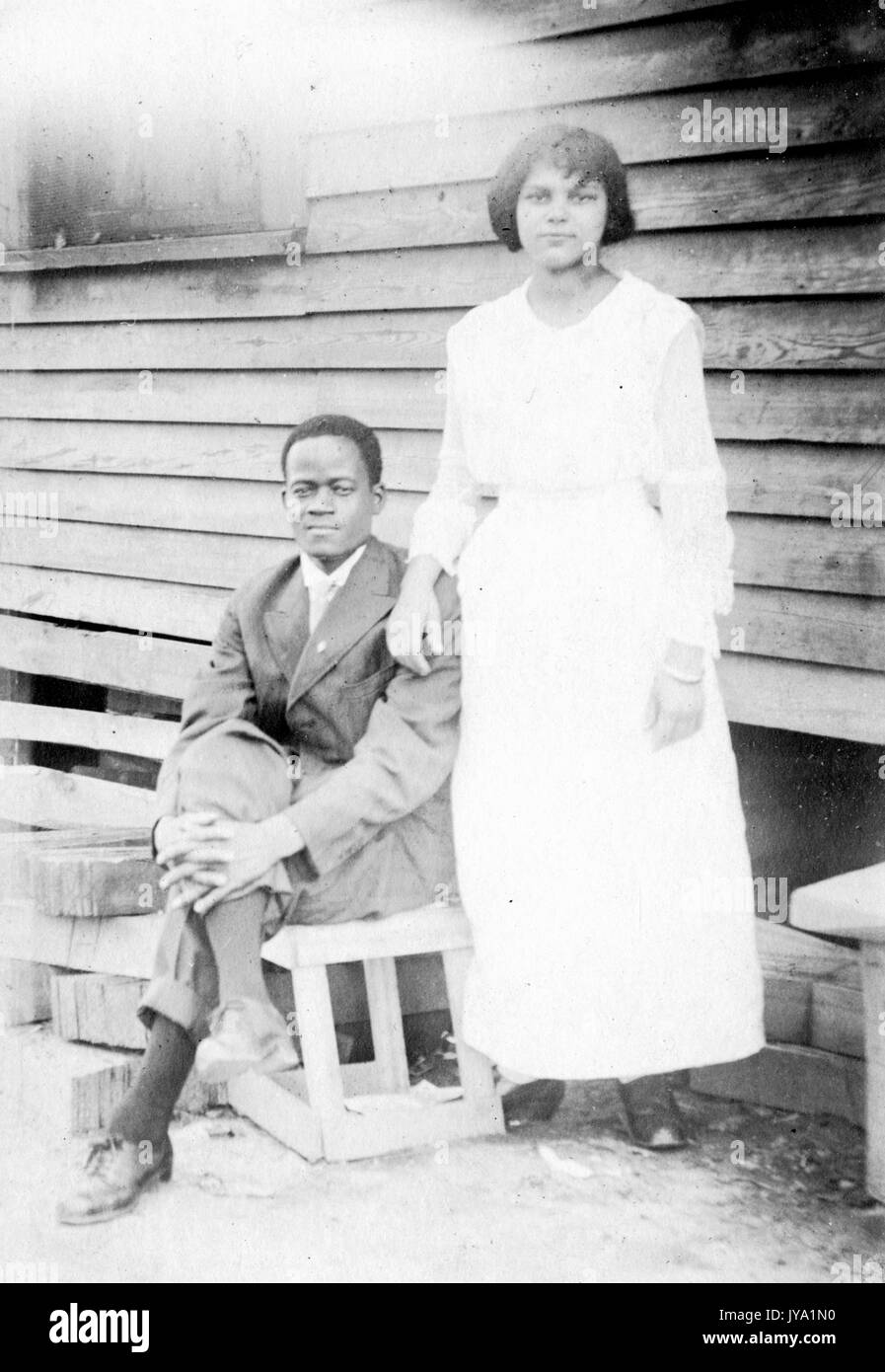 African-american couple posant devant une maison avec revêtement en bois, épouse wearing white dress et debout, mari assis sur une caisse en bois avec sa main sur son genou, 1910. Banque D'Images
