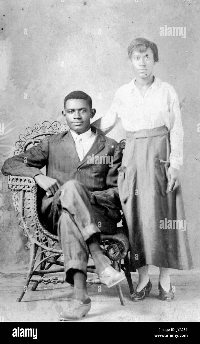 Portrait d'un couple afro-américain, l'homme porte un costume de couleur sombre et est assis dans une chaise en osier avec ses jambes croisées, la femme porte une chemise et une jupe de couleur claire et se tient à côté de lui avec son bras derrière son dos, 1920. Banque D'Images