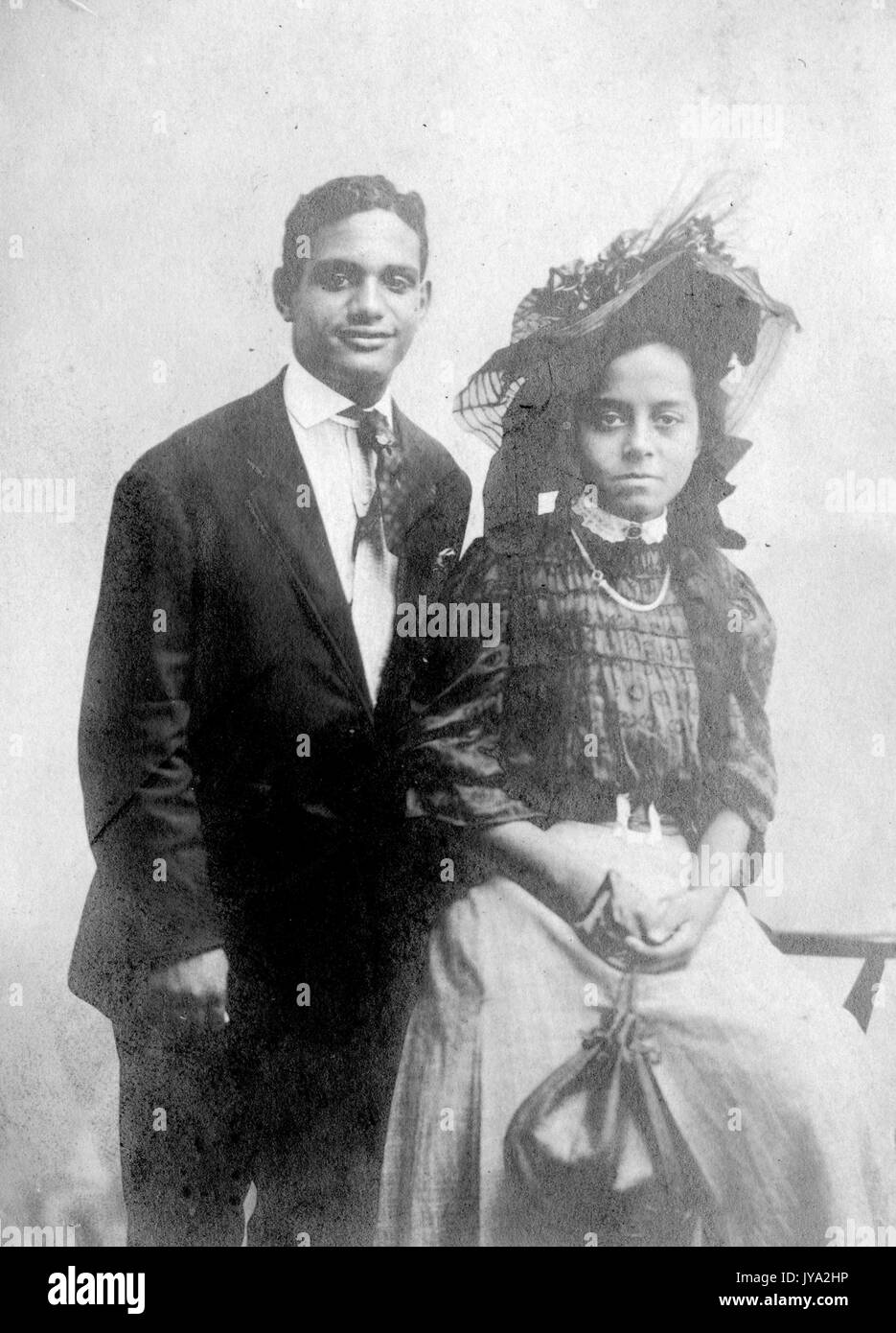 Couple afro-américain posant pour une photographie, l'homme portant un costume et souriant, les femmes assis sur une chaise devant l'homme, portant un chapeau, un chemisier et une jupe, avec une expression faciale sérieuse, Portsmouth, Virginie, 1920. Banque D'Images