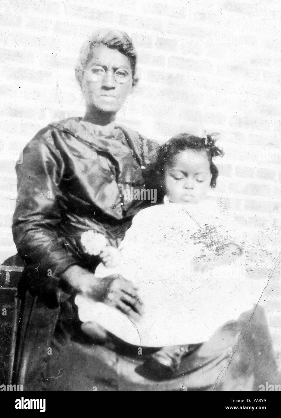 African American grand-mère et petit-fils, la grand-mère portant des lunettes avec ses cheveux en chignon, le jeune enfant assis sur ses genoux et vêtue d'une robe blanche avec ses yeux fermés, 1915. Banque D'Images