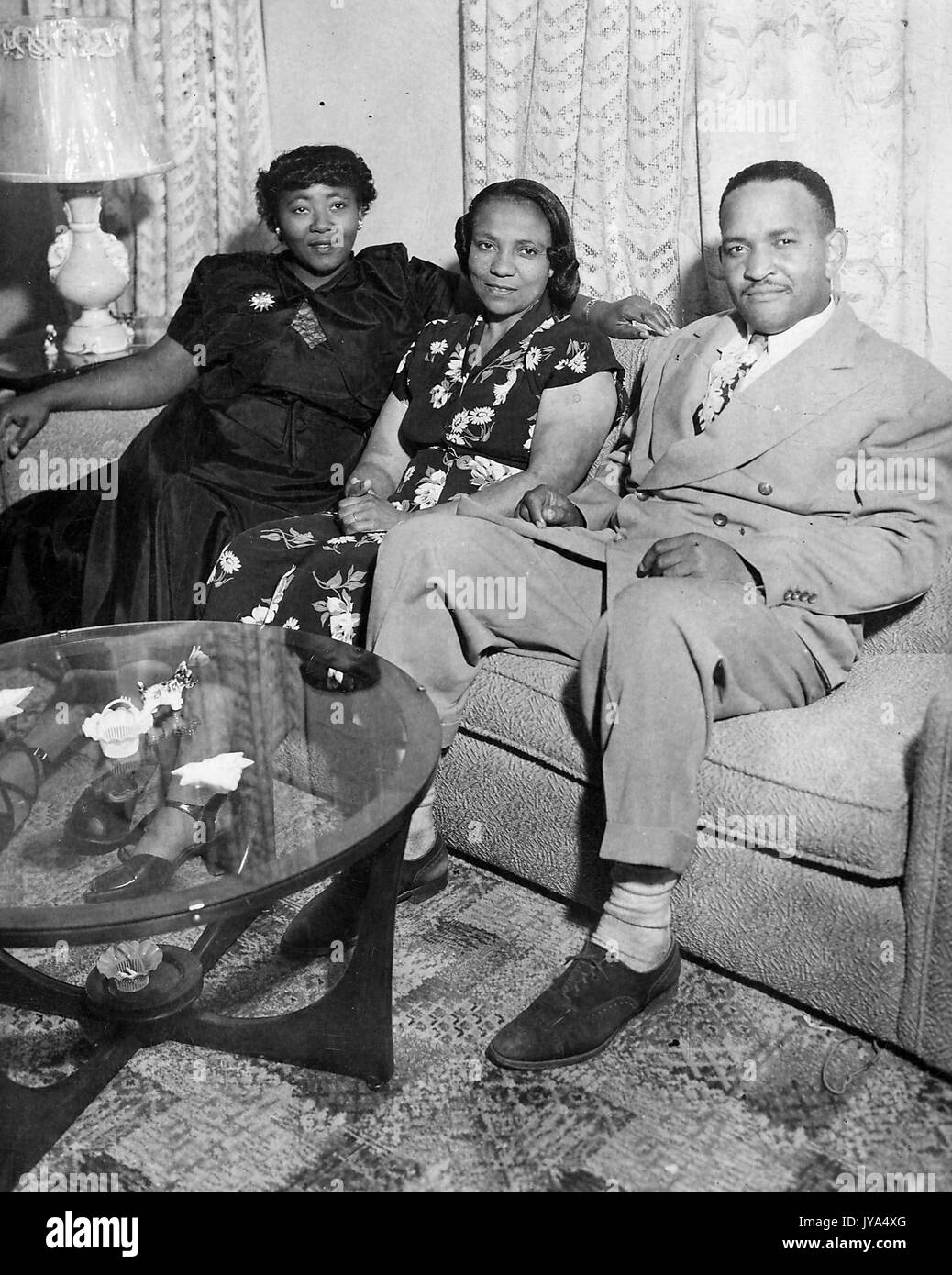 African-american family, père, mère et fille, assise sur un sofa dans leur maison, table basse en verre avec figurines visible dans l'avant-plan, une lampe et des rideaux à l'arrière-plan, 1960. Banque D'Images