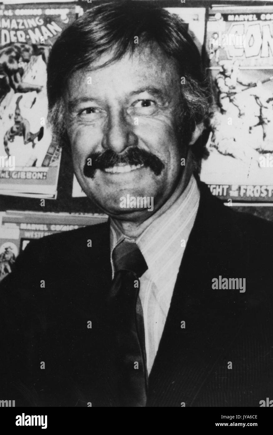 Portrait de l'auteur de comics, Stan Lee, portant un costume sombre et une cravate avec une chemise rayée, avec une expression du visage souriant, debout en face de nombreux comic books de sa création. 1975. Banque D'Images