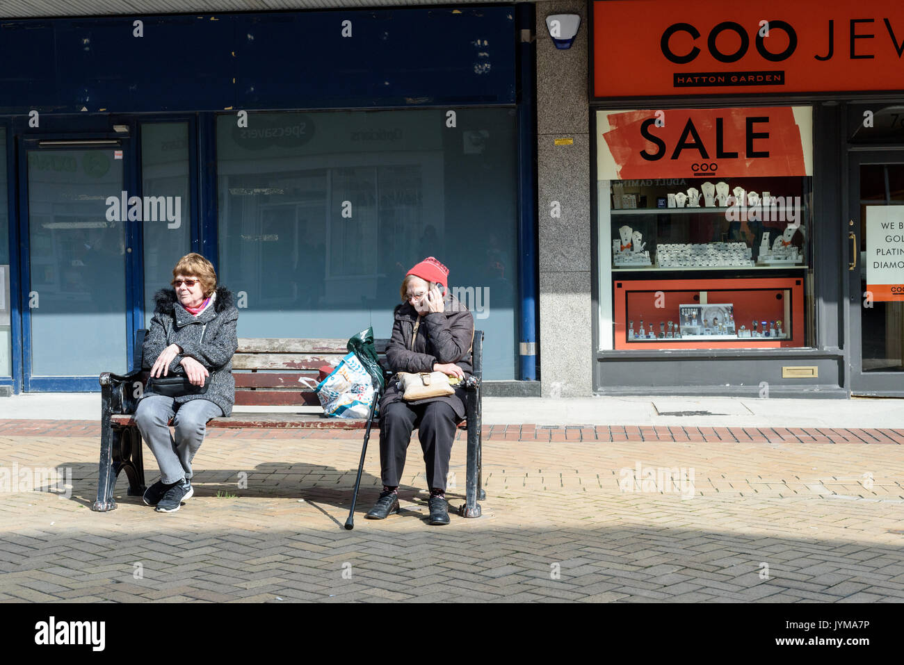 Deux vieilles femmes assises sur un banc public dans une rue piétonne pavée avec des magasins sont vides à l'arrière-plan Banque D'Images