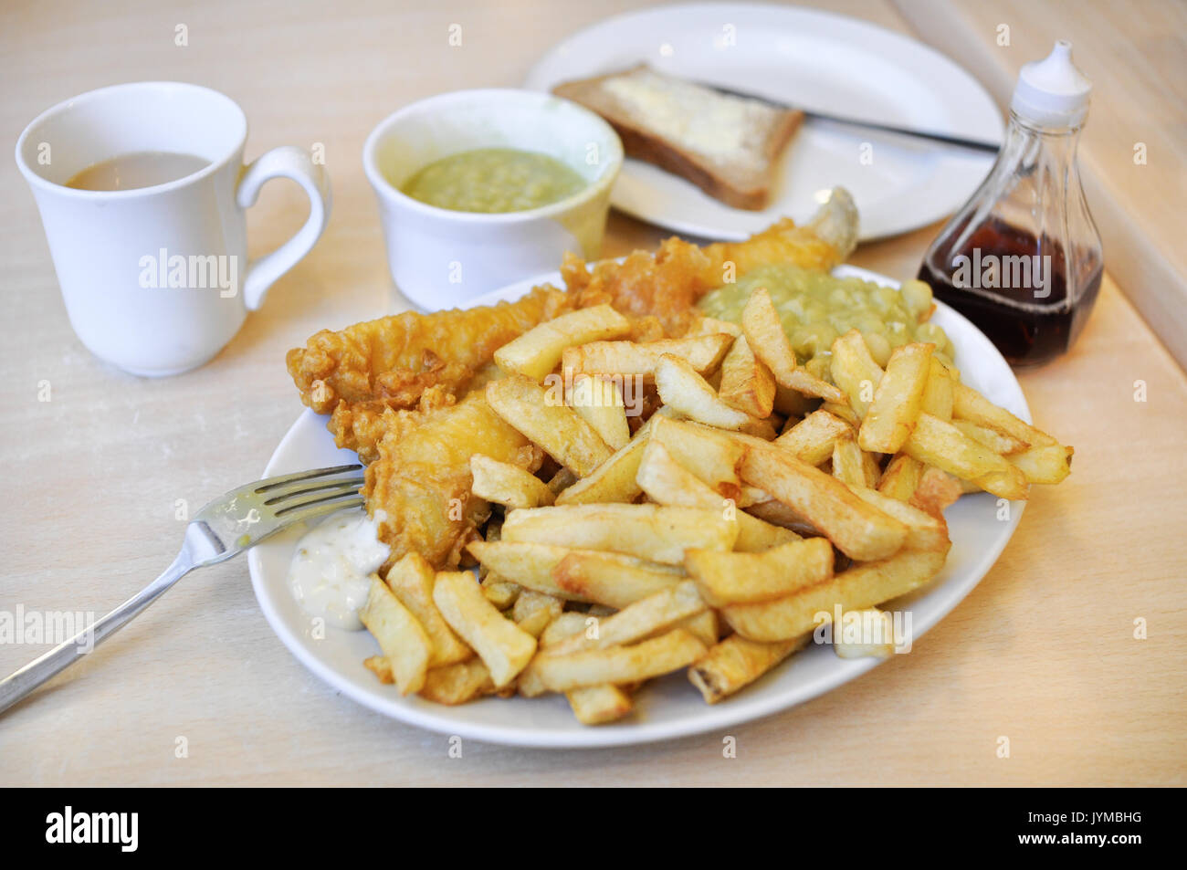Un plat de poisson et frites britannique traditionnel, avec des petits pois, du pain et du beurre, du vinaigre, de la sauce tartare, et une tasse de thé. Banque D'Images