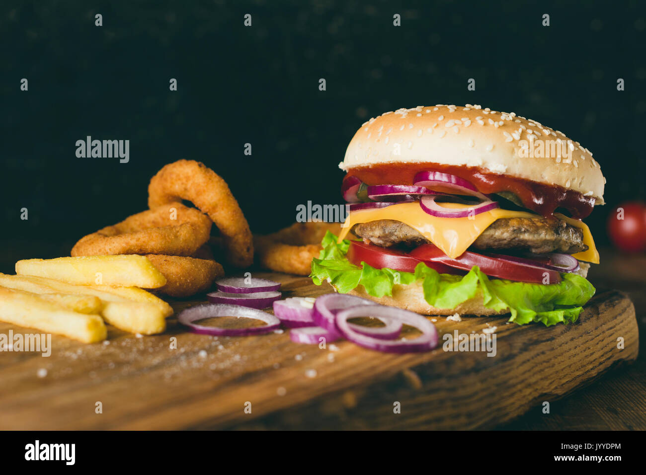 Cheeseburger, frites et oignons sur planche à découper en bois sur fond de bois. Vue rapprochée, selective focus. Concept de restauration rapide Banque D'Images