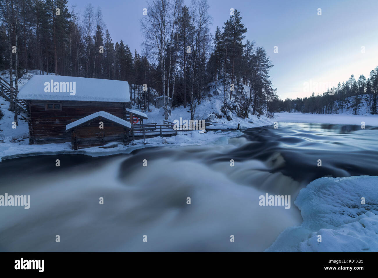 Une cabane en bois entouré par la rivière rapide et Snowy Woods au crépuscule Juuma Myllykoski région Laponie Finlande Europe Banque D'Images