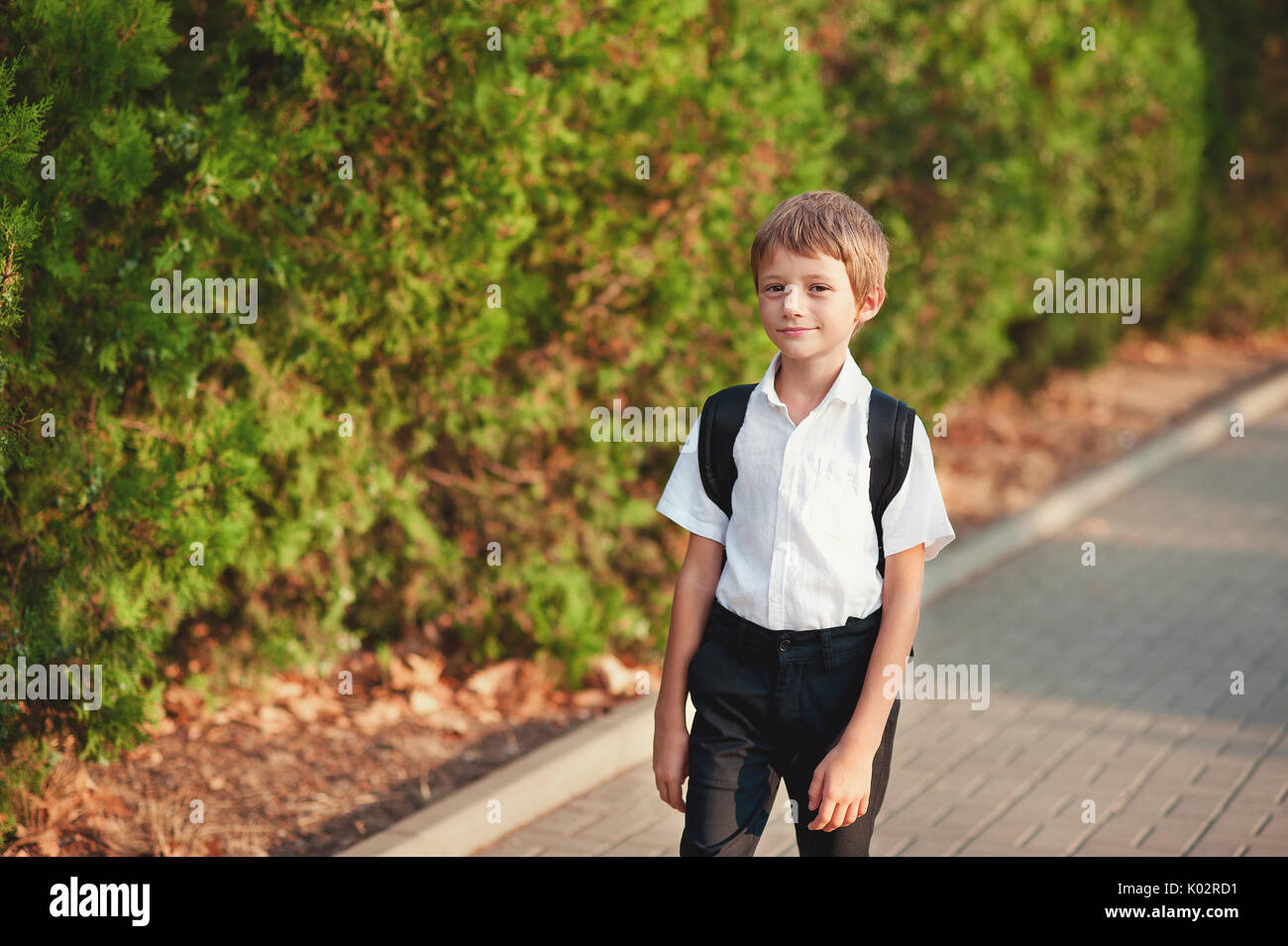 Petit écolier revient de l'école dans la bonne humeur. Le garçon va sur un sentier. Il a soulevé les yeux au ciel et sourit. Derrière les épaules à l'élève une besace. Banque D'Images