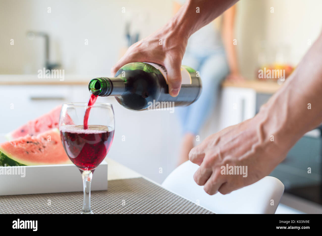 Mari de verser un verre de vin rouge pour sa femme qui est assis sur le comptoir de la cuisine derrière lui. La main, bouteille de vin et le verre close up Banque D'Images