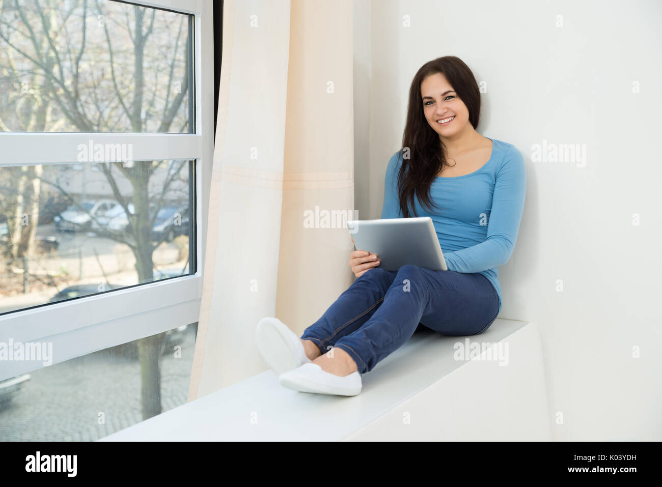 Portrait Of A Smiling Young Woman Sitting Near Window Using Digital Tablet Banque D'Images