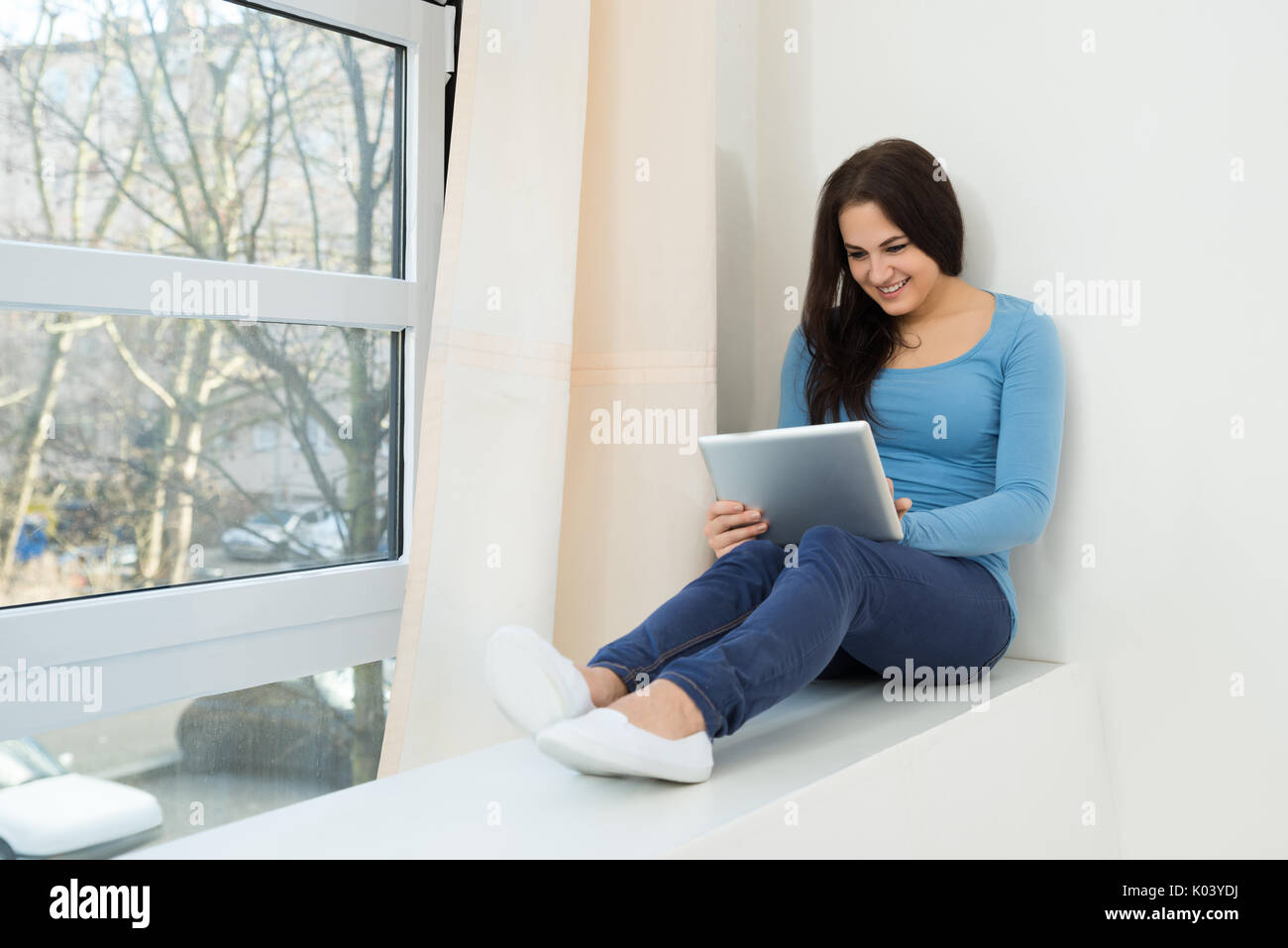 Portrait Of A Smiling Young Woman Sitting Near Window Using Digital Tablet Banque D'Images