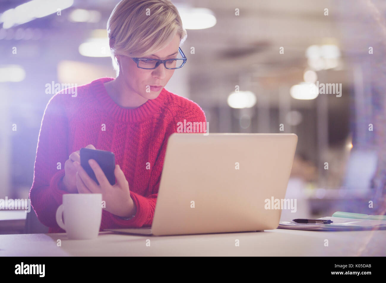 Femme sérieuse travaillant tard, using cell phone at desk in office Banque D'Images