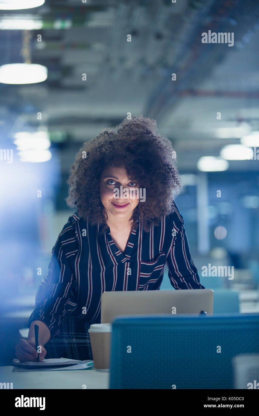 Portrait of smiling confident businesswoman travailler tard au bureau sombre dans l'ordinateur portable Banque D'Images
