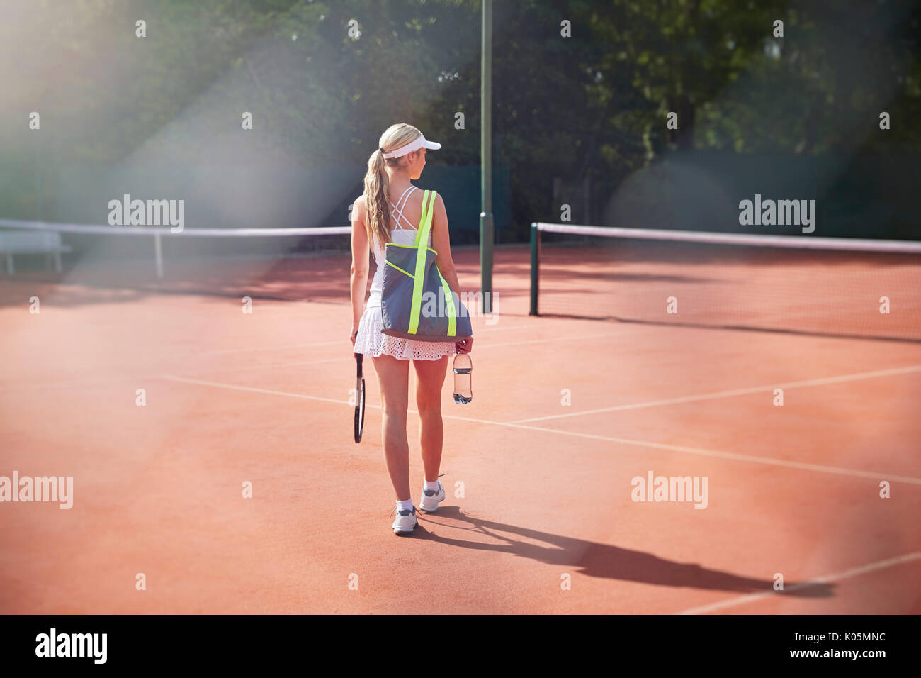 Les jeunes tennis player marche avec raquette de tennis, un sac et une bouteille d'eau sur terre battue ensoleillée Banque D'Images