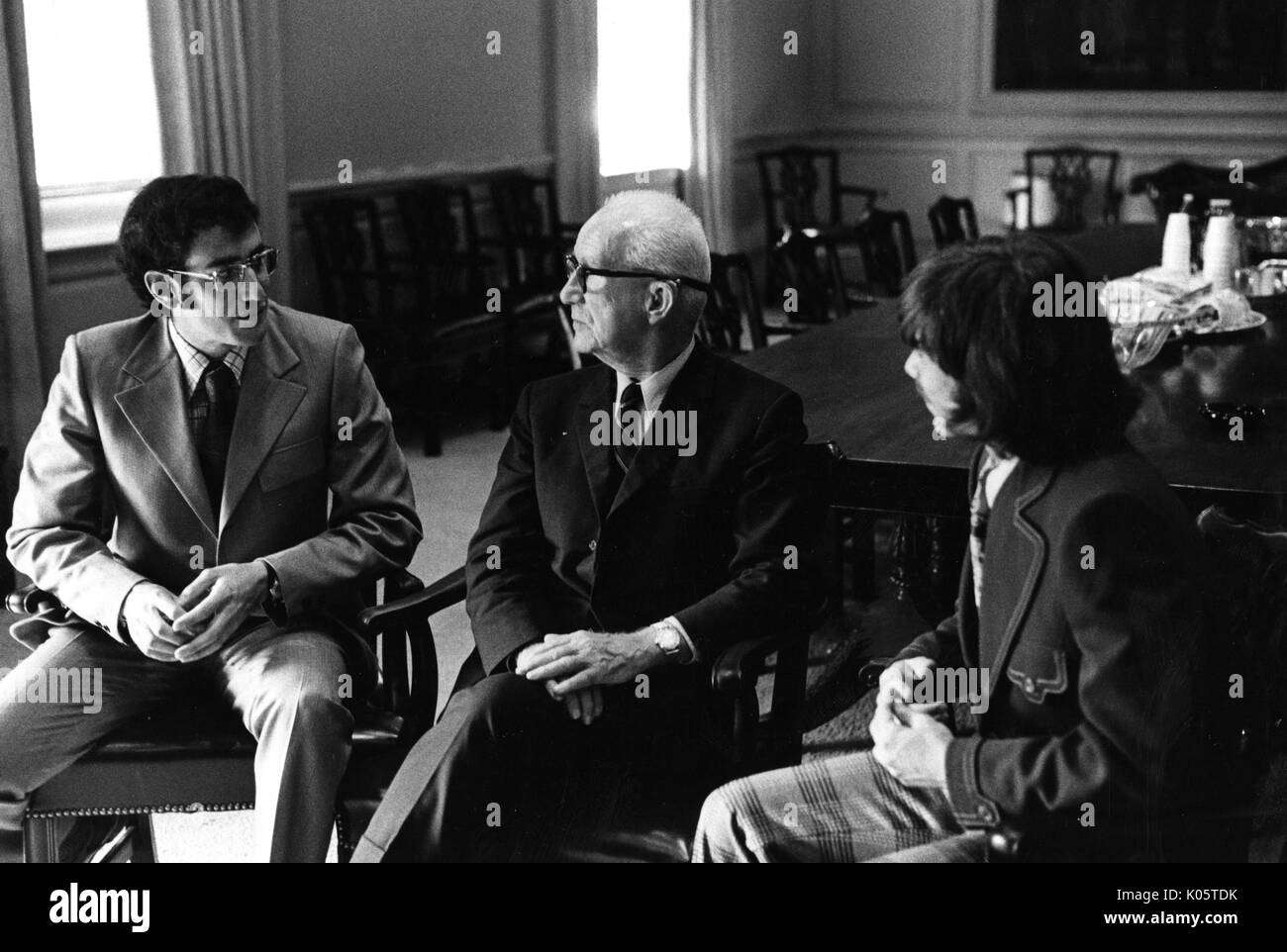 Portrait de trois-quarts de l'architecte Buckminster Fuller, assis dans une chaise en bois entre deux autres hommes, Fuller portant des lunettes et un costume sombre et une chemise blanche avec une cravate rayée, en face de l'homme à sa droite, avec ses mains jointes sur ses genoux, avec un sérieux de l'expression du visage. 1970. Banque D'Images