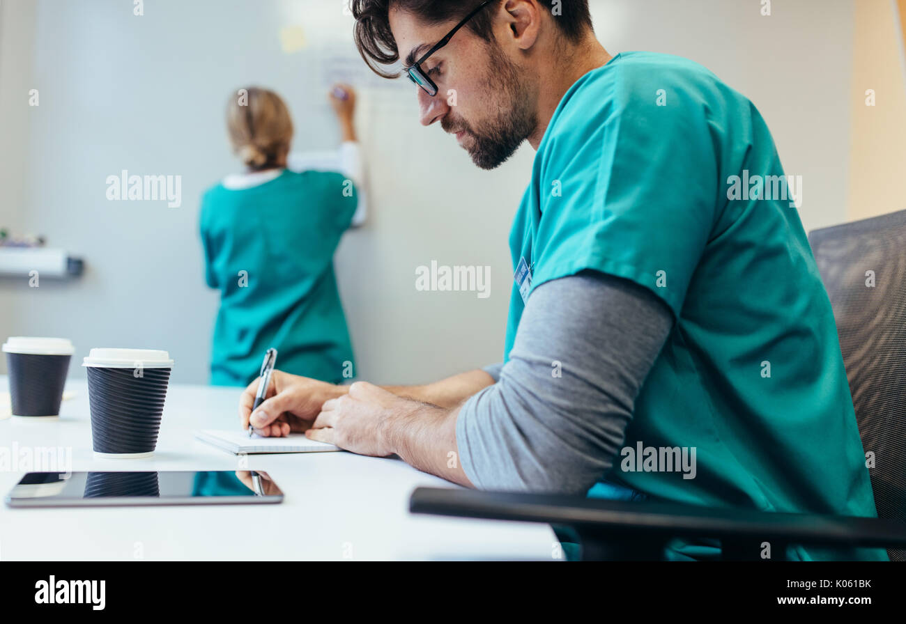 Vue latérale du travailleur de la santé assis dans la salle de l'hôpital notes d'écriture au féminin en arrière-plan. Banque D'Images