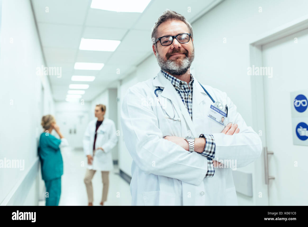 Portrait of positive doctor standing in hospital hallway, les bras croisés. Médecin mature avec des collègues parler en arrière-plan. Banque D'Images