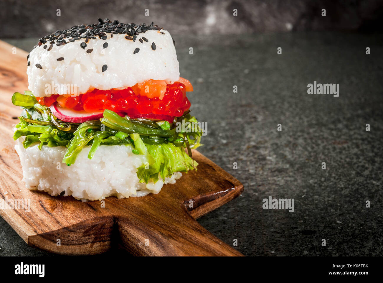 L'alimentation hybride de tendance. Japanese cuisine asiatique. Sushi-burger, sandwich au saumon, wakame hayashi, daikon, gingembre, caviar rouge. Table en pierre noire, avec du soja Banque D'Images