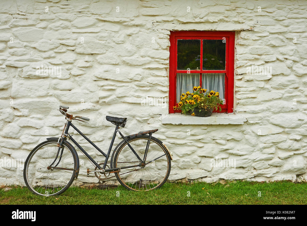 Un vieux vélo appuyé contre un bâtiment en pierre de chaux lavée avec une fenêtre peinte rouge et jaune fleurs Banque D'Images