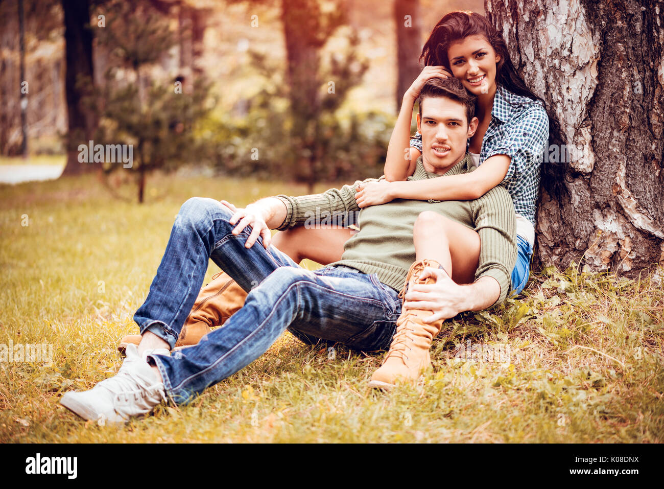 Belle lovely couple assis sur l'herbe à côté de l'arbre et bénéficiant à sunny park en couleurs de l'automne. looking at camera. Banque D'Images