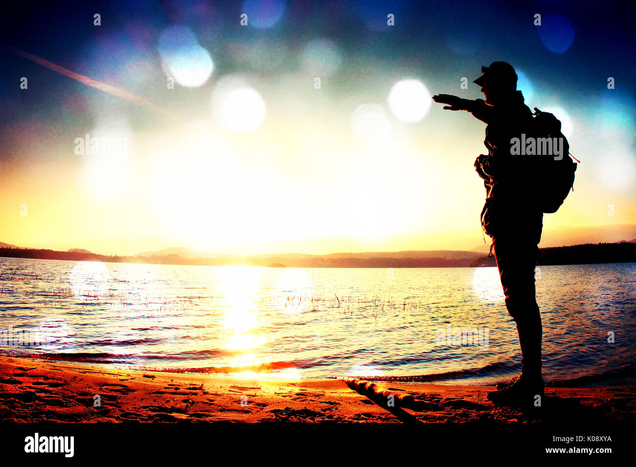 L'effet de grain de film. Homme heureux avec la main dans l'air. Grand randonneur dans les vêtements de sport sac à dos sombre avec des stands sur la plage admirer le coucher du soleil à l'horizon avec l'axe Banque D'Images