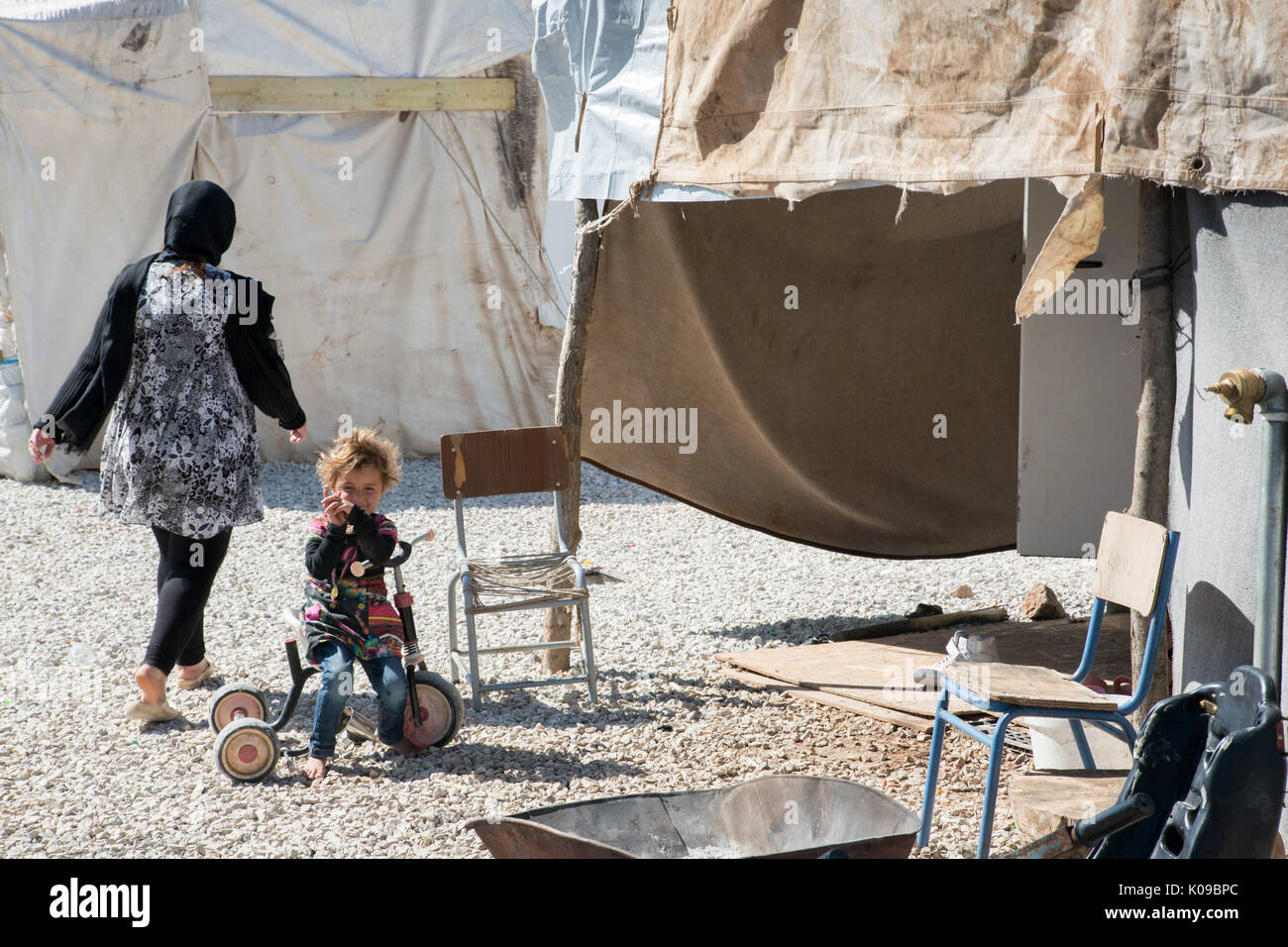 Au camp Ritsona en Grèce pour les réfugiés syriens, un enfant aux pieds nus sur un tricycle regarde la caméra comme une femme adulte ( retour à la caméra) s'éloigne. Banque D'Images