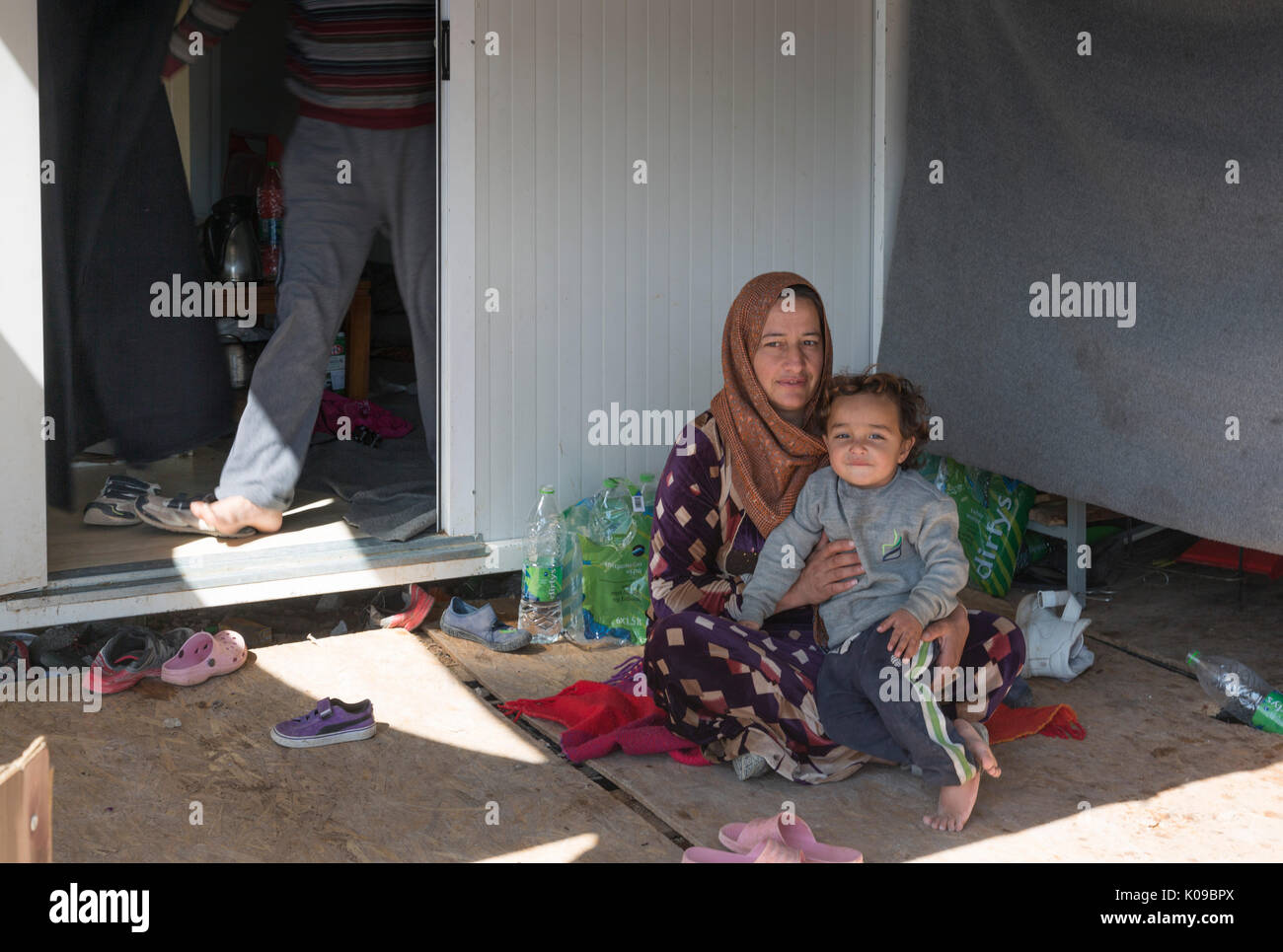 Un réfugié mère se repose à l'ombre de sa maison usinée dans le camp de réfugiés syriens Ritsona en Grèce. Les jambes de son mari dans la porte derrière. Banque D'Images