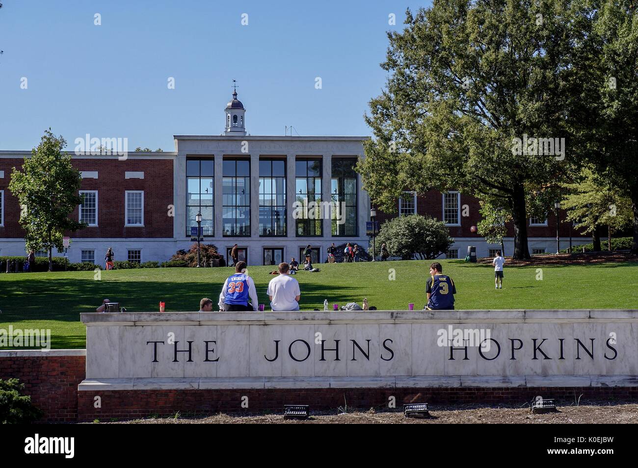 Vue depuis la rue Charles de la « plage » et la bibliothèque Milton S Eisenhower de l'université Johns Hopkins ; trois étudiants de sexe masculin, face à la caméra, s'assoient sur le panneau de l'université en marbre, d'autres étudiants se mettent à bronzer et jouent au catch sur la colline herbeuse ; La tour de l'horloge de Gilman Hall, un bâtiment des humanités, surplombe le sommet de la bibliothèque; Baltimore, Maryland, mars 2014. Avec la permission d'Eric Chen. Banque D'Images