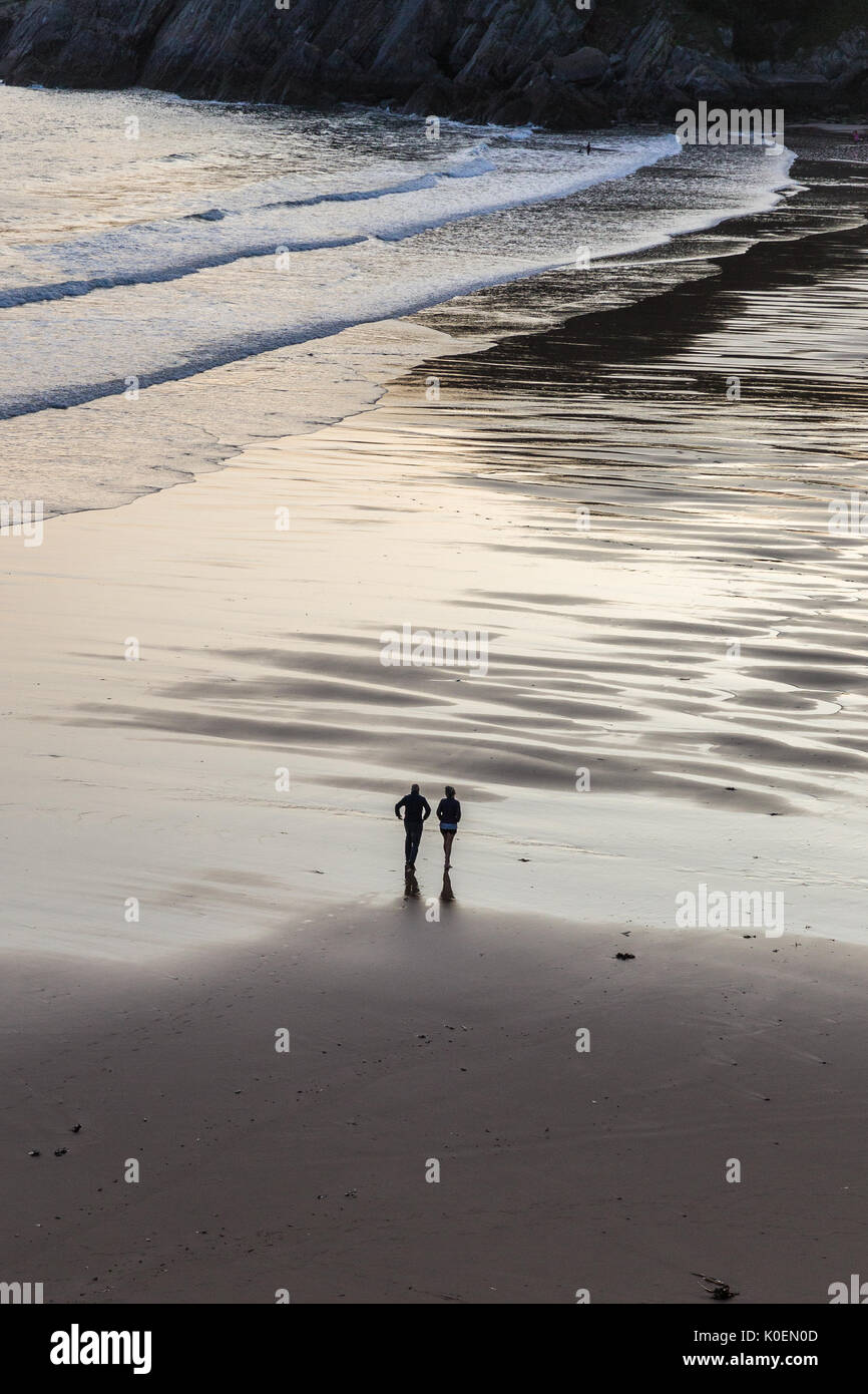 Une scène côtière avec deux chiffres silloutted traversant le sable humide au coucher du soleil sur Caswell Bay sur la péninsule de Gower au Pays de Galles, Royaume-Uni Banque D'Images