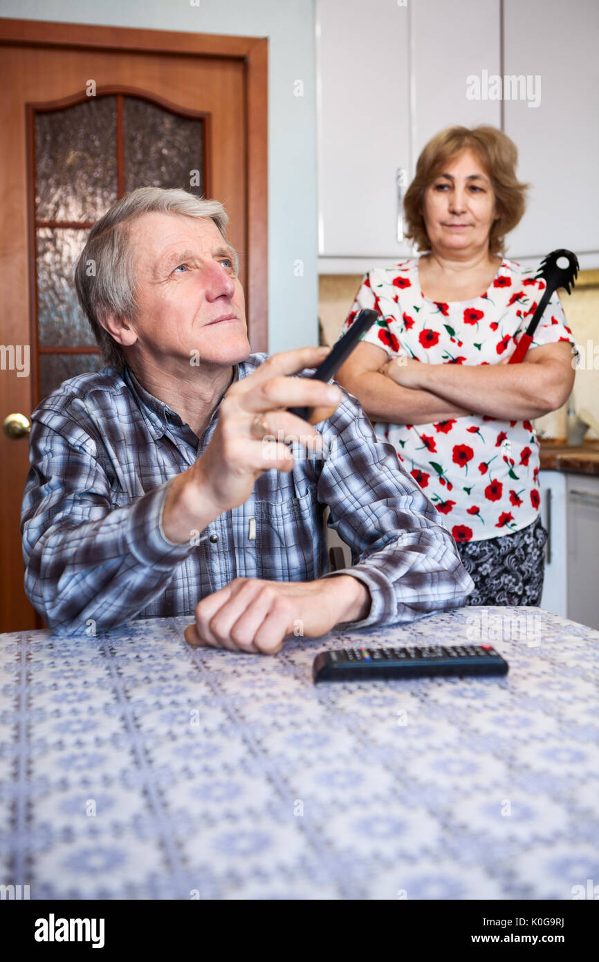Relations de couple d'Europe senior, l'homme regardant la TV avec télécommande tandis que son épouse en colère debout derrière dans la cuisine Banque D'Images