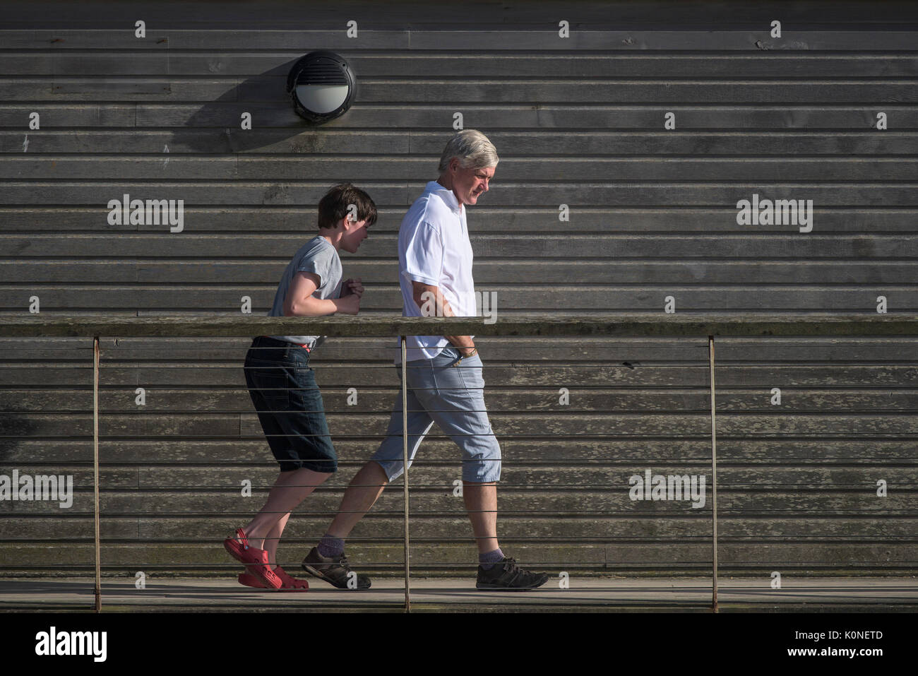 Un homme marche sur une passerelle dans le vent avec son fils à l'abri lui behibd. Banque D'Images