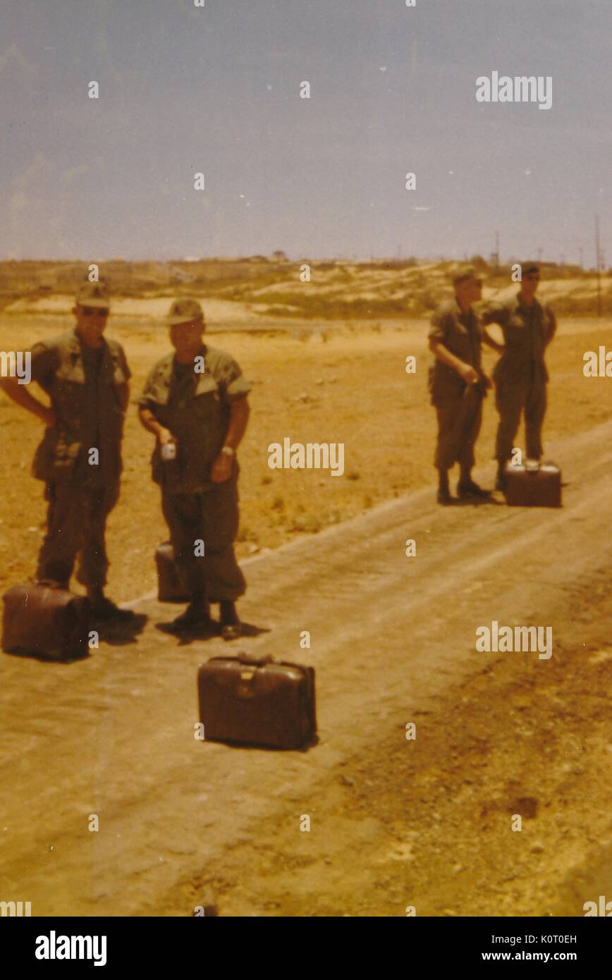Un groupe de quatre soldats faire la conversation pendant qu'ils attendent, avec leurs valises, pour leur transport à arriver, Rocky, avec petit terrain poussiéreux de la végétation dans l'arrière-plan, Vietnam, 1964. Banque D'Images