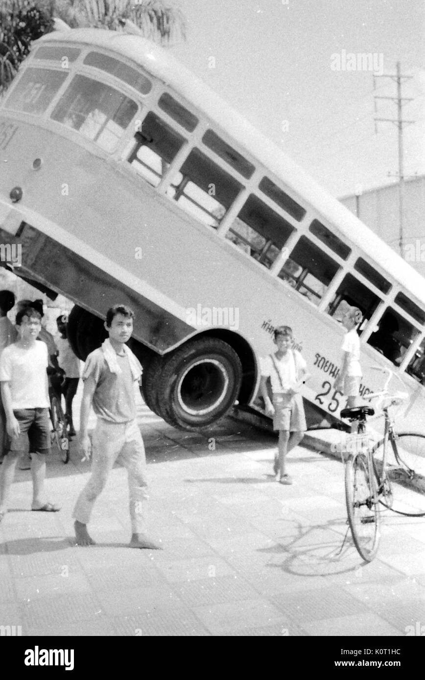 Se penchant sur le bus de bord d'un pont, dans un cadre urbain, les pneus arrière en l'air, avec une foule d'hommes et garçons rassembler autour de l'autobus, Vietnam, 1969. Banque D'Images