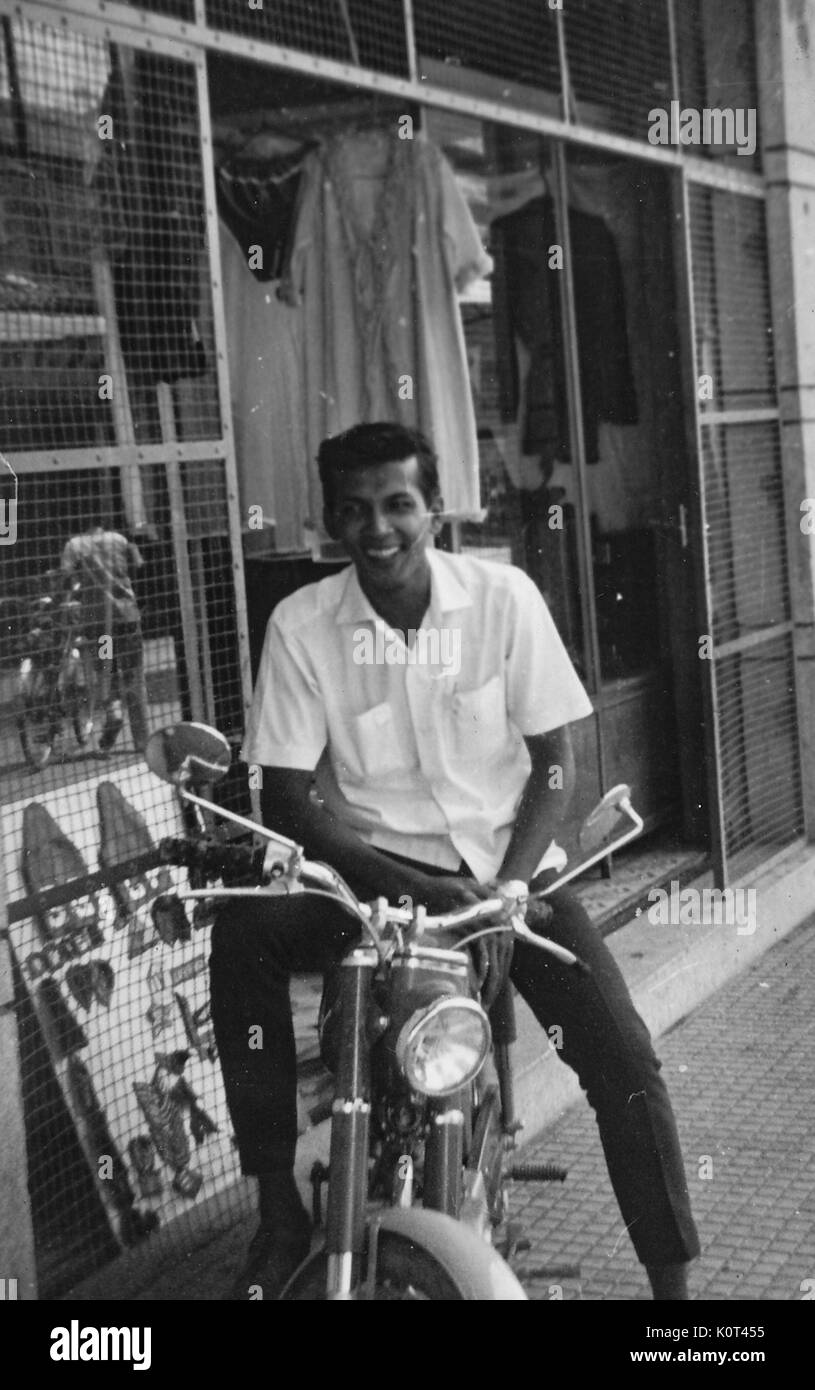 A smiling man vietnamien assis sur une moto qui est garé sur le trottoir en face d'un magasin de vente au détail, Vietnam, 1967. Banque D'Images