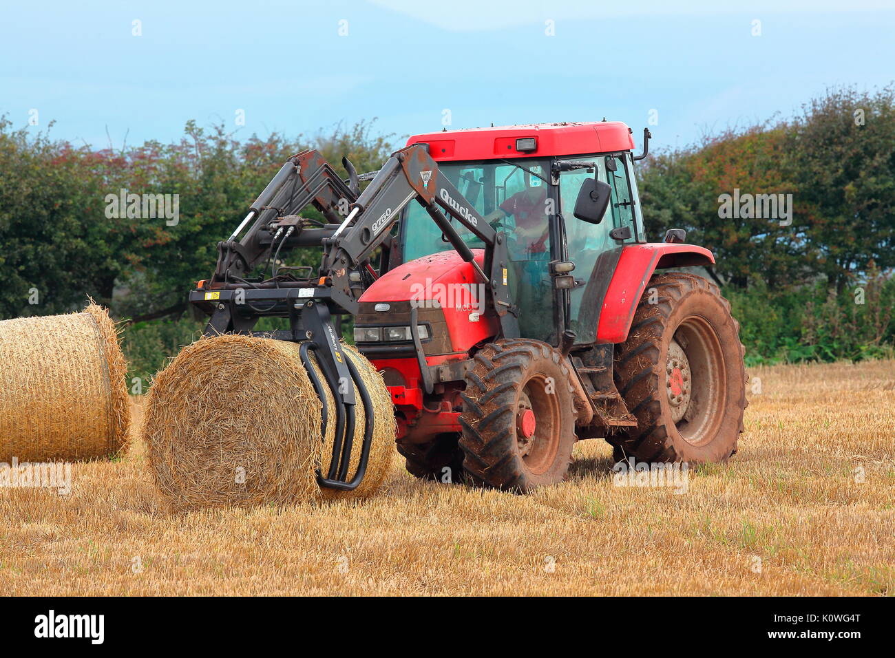 Un agriculteur perçoit sa série de bottes de paille dans un champ avec son tracteur spécialement adaptés avec une pince pour ramasser les balles énormes et les faisant tourner. Banque D'Images