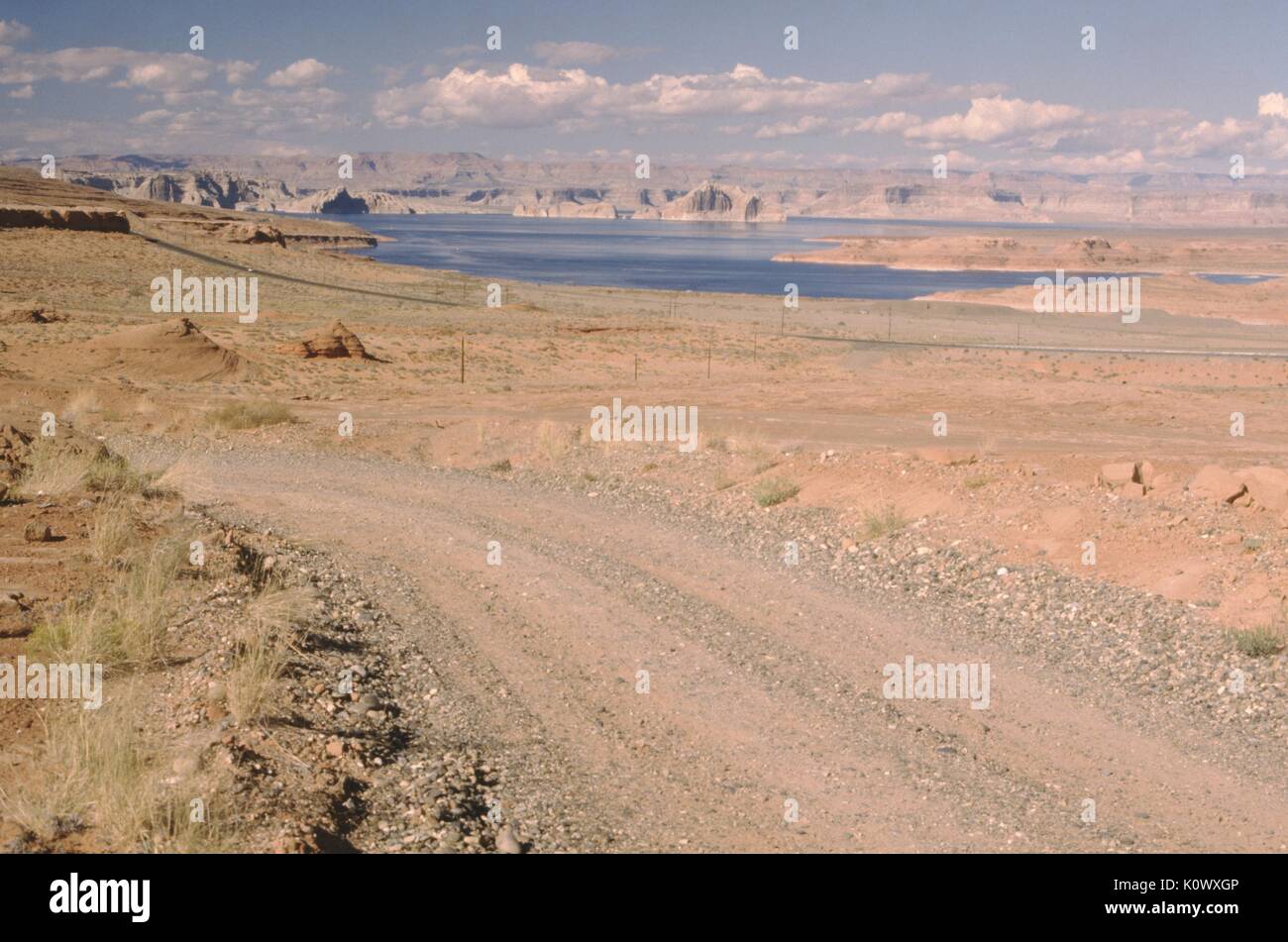 Route de gravier à travers un paysage désertique, des sols d'argile rouge entourant la route, mesas menant jusqu'à une étendue d'eau visible à l'arrière-plan, une couverture nuageuse basse, journée ensoleillée, 1969. Crédit photo Smith Collection/Gado/Getty Images. Banque D'Images