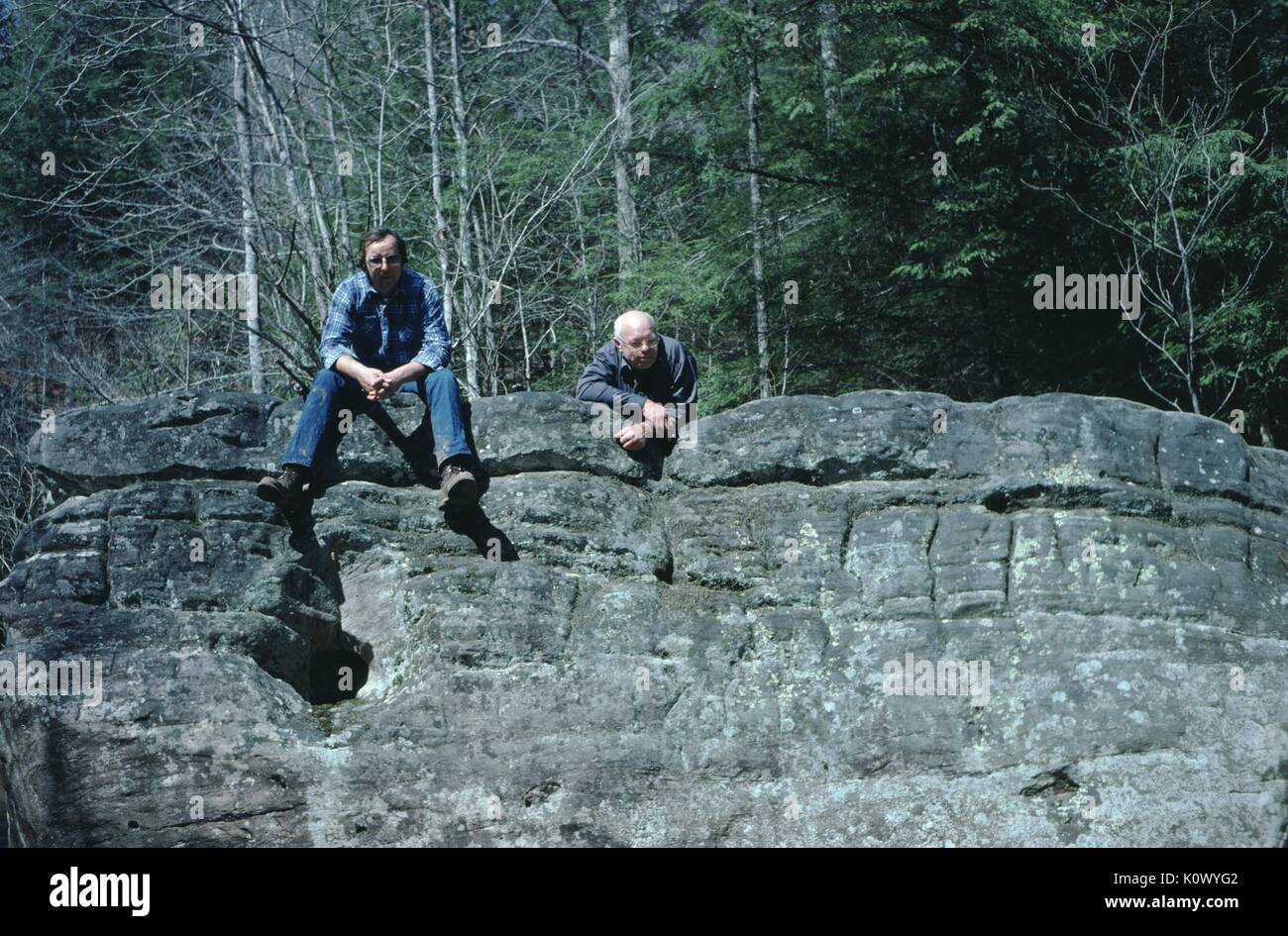Deux hommes dans les bois, sur le bord d'un affleurement rocheux, un homme assis sur les rochers, les jambes suspendues sur le bord et ses mains croisées, l'autre penché sur les rochers, du grain rôti Creek, Kentucky, 1975. Crédit photo Smith Collection/Gado/Getty Images. Banque D'Images