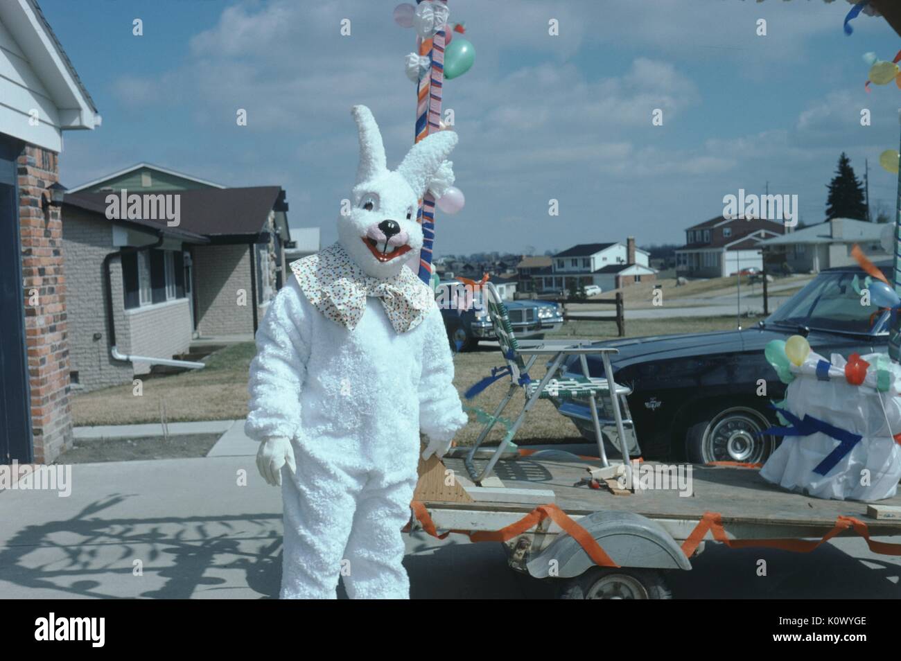 Mascot dans un grand costume de lapin blanc, debout dans l'allée d'une maison de banlieue, une chaise longue sur une remorque à plateau avec des banderoles et des ballons, avant une parade, 1975. Crédit photo Smith Collection/Gado/Getty Images. Banque D'Images