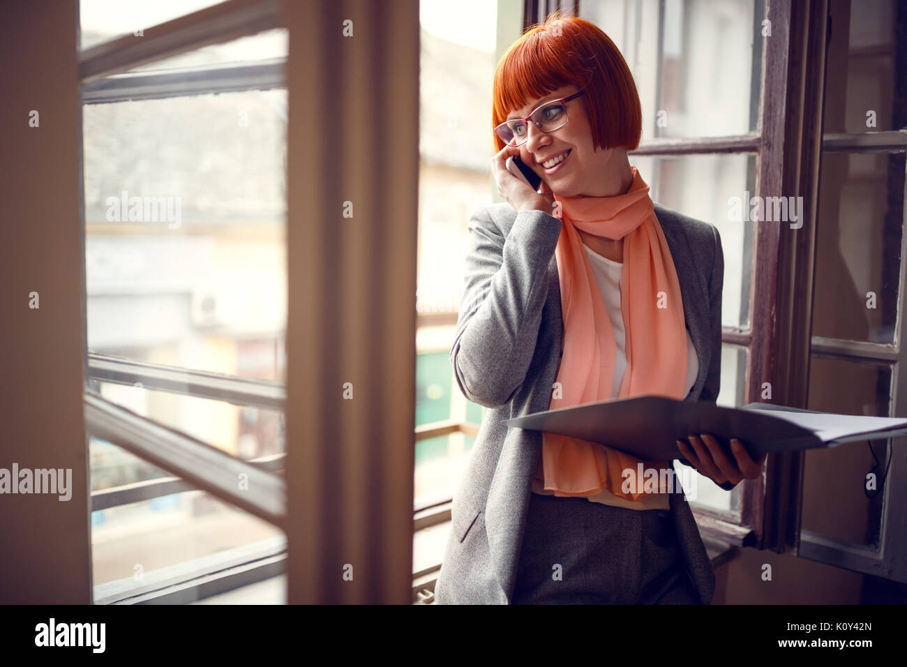 Jeune femme par fenêtre dans l'organisation de réunions de bureau avec téléphone cellulaire Banque D'Images