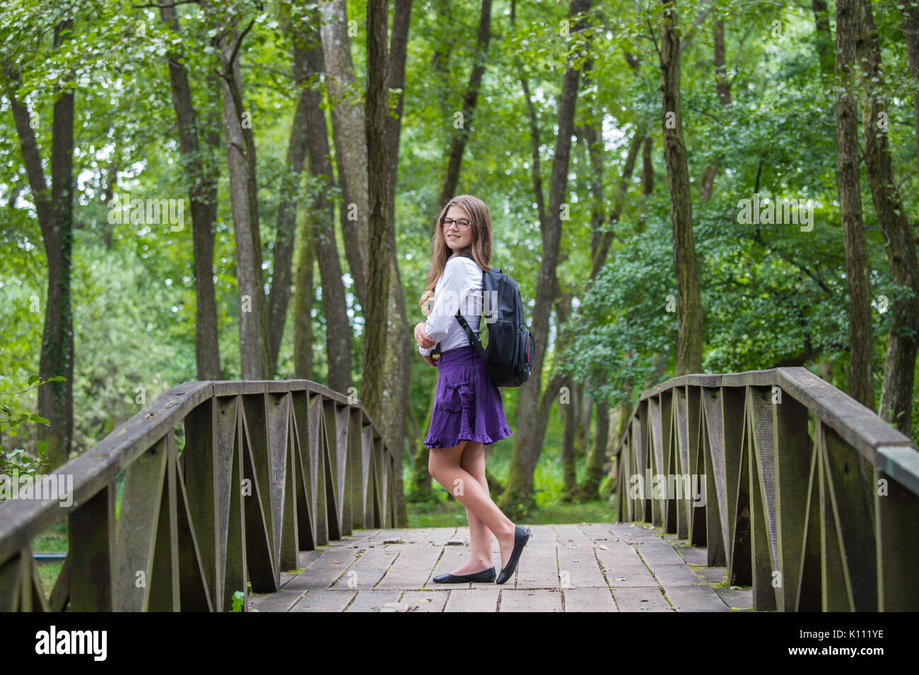 Belle belle blonde fille de l'école d'enfant souriant avec des lunettes, une chemise blanche, jupe violet et noir sac à dos debout sur un pont en bois dans la nature tre Banque D'Images